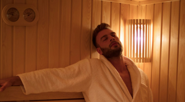 A man sits in a sauna wearing a white robe, surrounded by wooden walls