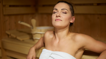 A woman sits in a sauna with her eyes closed, relaxing and soaking in the heat. She is wrapped in a white towel, enjoying the peaceful atmosphere.