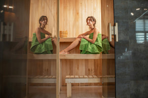 A women sits in a sauna, wrapped in a green towel surrounded by wooden walls.